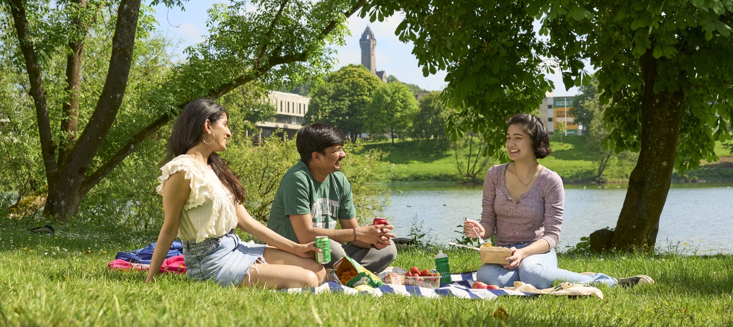 Students relaxing on the grass, having a picnic with Airthrey Loch and Wallace monument in the background.
