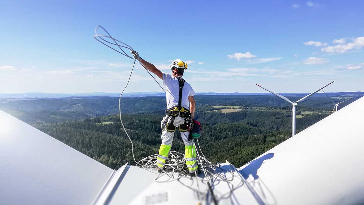 Professional rope access technician standing on roof (hub) of wind turbine and pulling rope up. Sun is behind wind turbine.