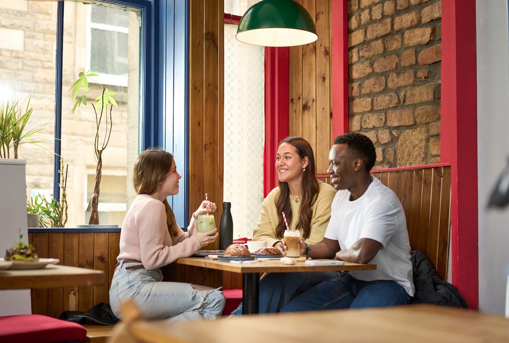 Three friends enjoy drinks and snacks together at a cozy café booth with colorful wooden and brick walls.