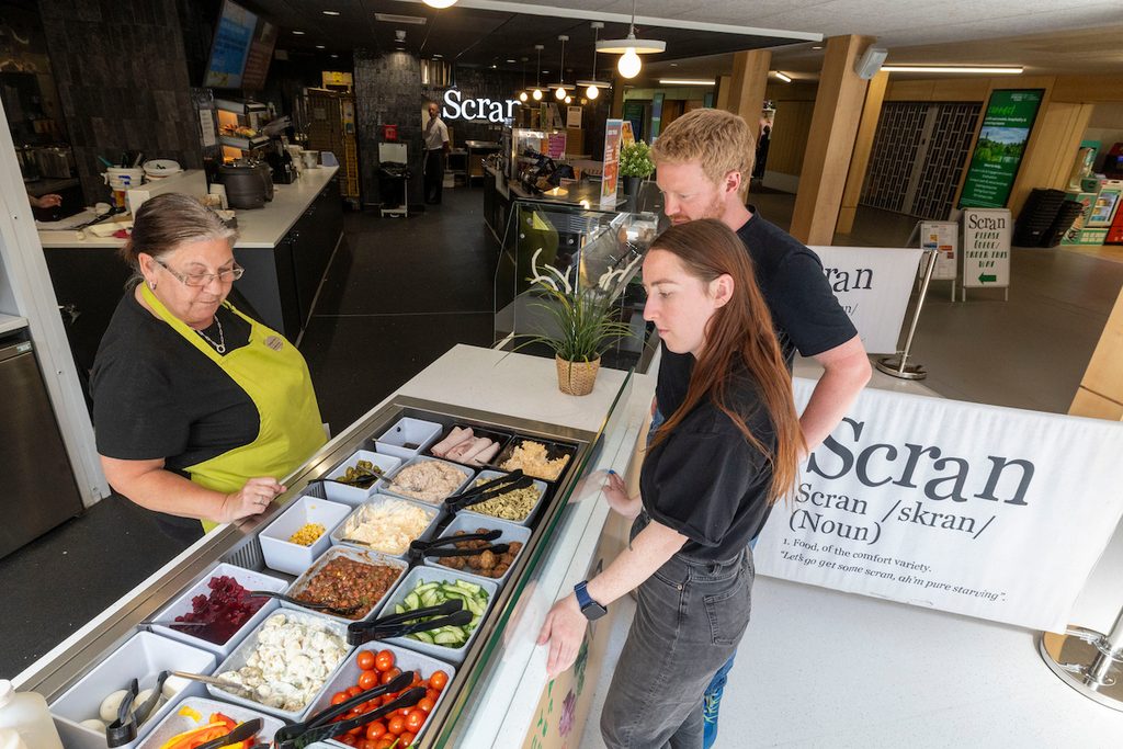Two individuals select food from a self-serve counter at a café called "Scran," while a staff member assists.