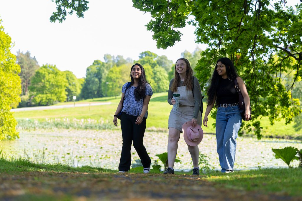 Three individuals stroll along a sunlit path beside a pond, smiling and enjoying a pleasant day outdoors.