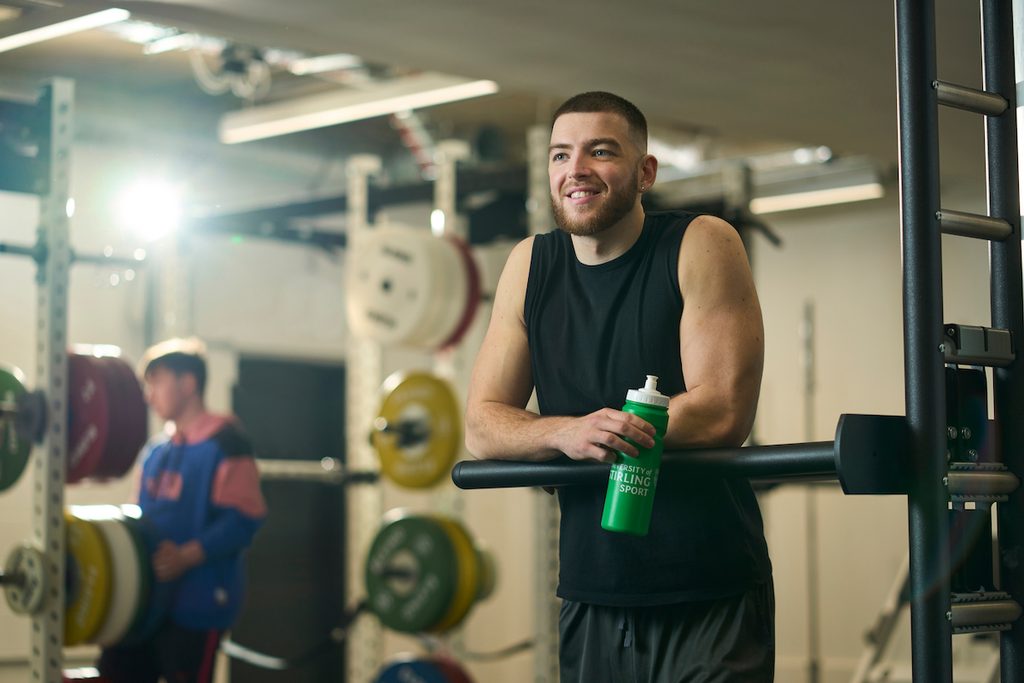 A person with a beard smiles while holding a green water bottle in a gym, standing near weightlifting equipment.